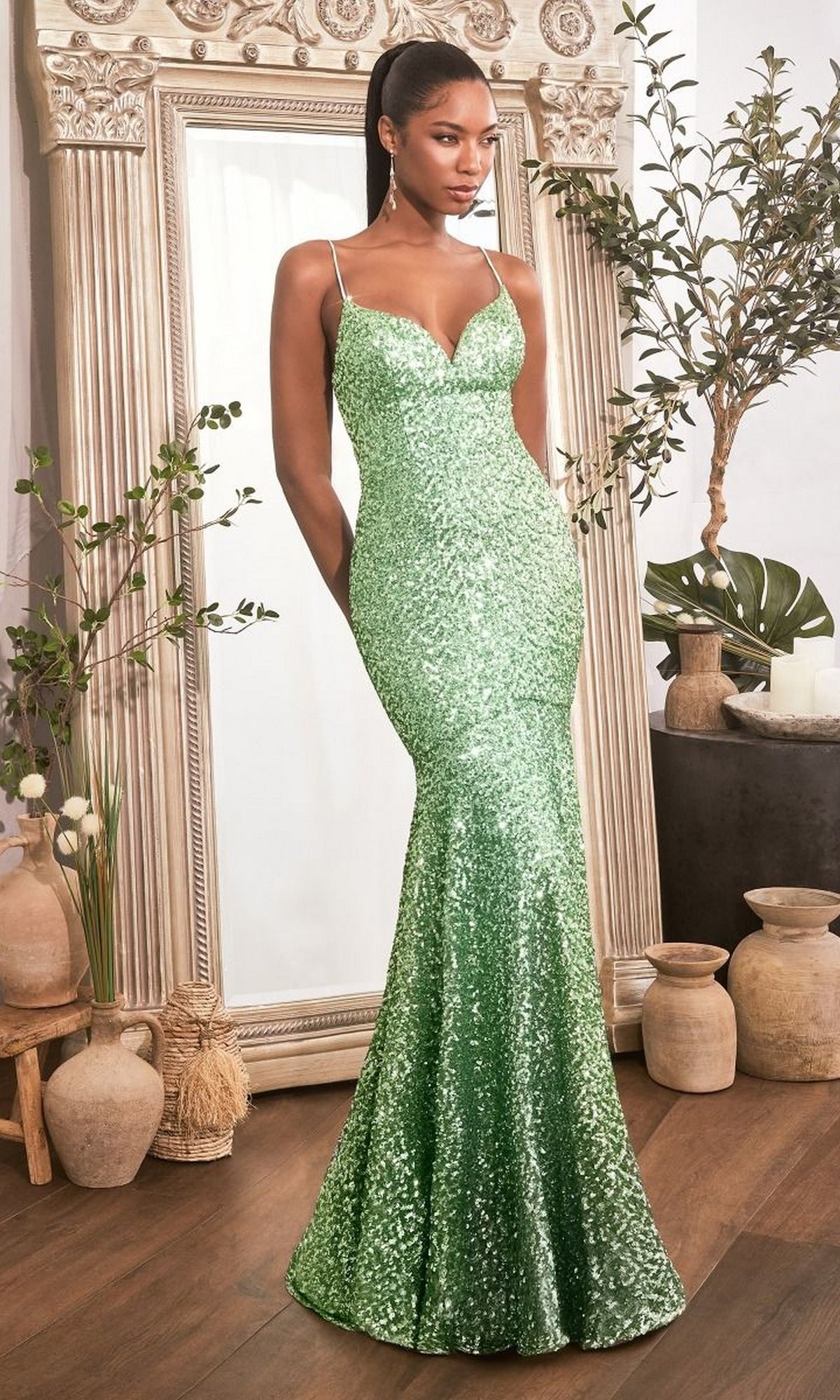 A woman models the Lace-Up Open-Back Long Sequin Prom Dress CH066, a fitted sparkly green gown, standing before a large mirror surrounded by decorative pottery and plants in a well-lit room.